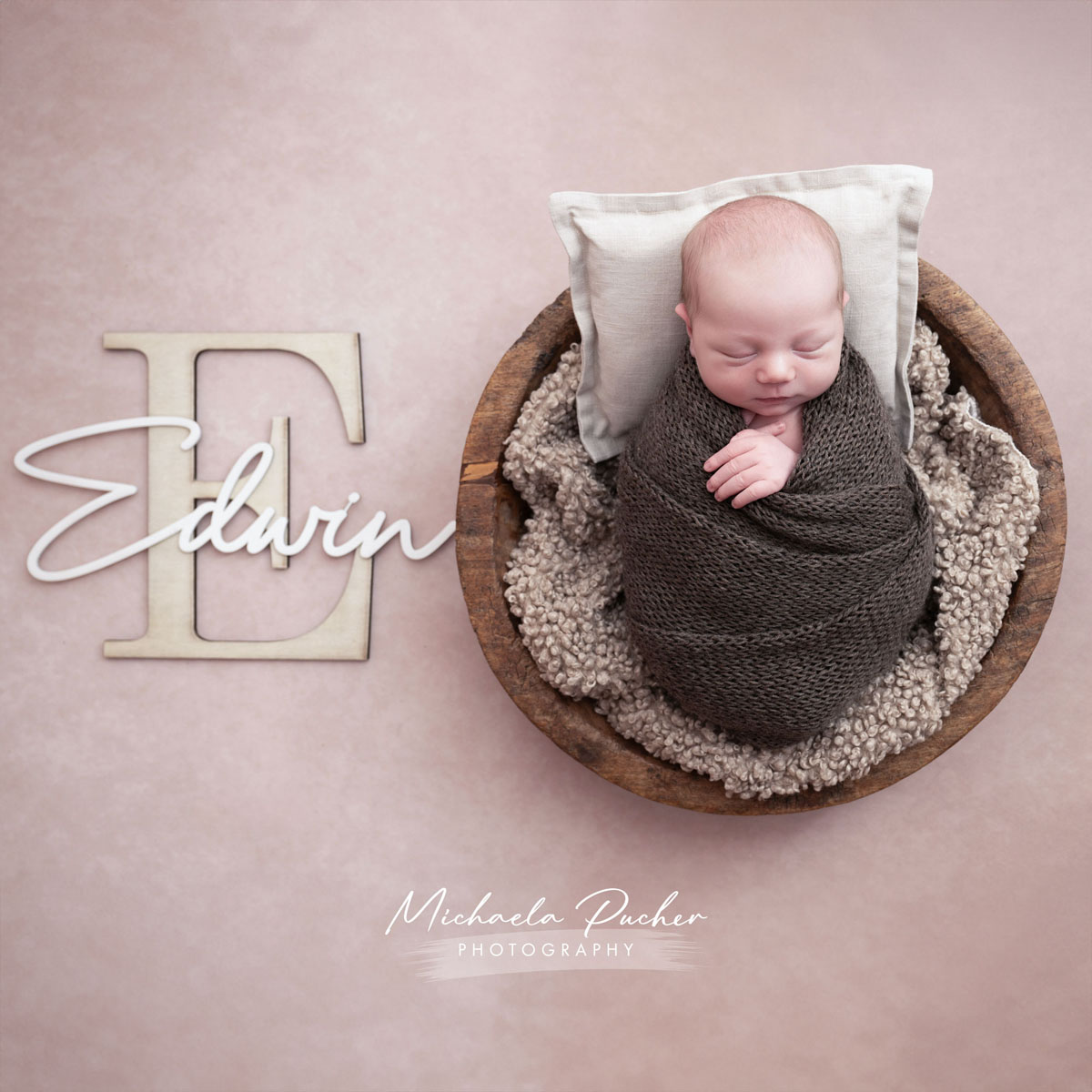 Schlafendes Neugeborenes liegt eingewickelt in dunklem Stricktuch in einer Holzschale; daneben steht der Name „Edwin“ mit großem Anfangsbuchstaben E auf beigefarbenem Hintergrund.